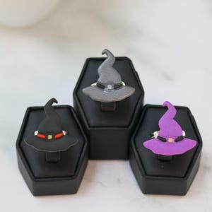 May include: Three Halloween-themed rings, each featuring a witch hat design. The rings are displayed on black stands. One ring is black with a red band, another is gray, and the third is purple. The hats have a silver band and buckle.