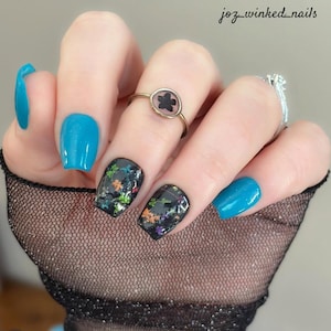 May include: Close-up of a hand with blue and black nail polish. The black nails have a glittery design with colorful accents. The hand is wearing a ring with a black flower design.