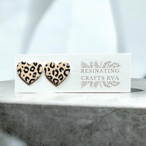 May include: A pair of heart-shaped earrings with a leopard print design. The earrings are made of resin and are a light beige color with black spots. The earrings are shown on a white card with the text "RESINATING CRAFTS RVA".