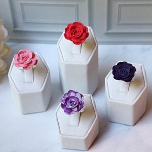 May include: Four flower rings in red, pink, purple, and dark purple are displayed on white stands. Each ring features a detailed floral design. The rings are set against a neutral background, highlighting the vibrant colors of the flowers.