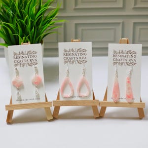 May include: Three pairs of handmade earrings displayed on small wooden easels. The earrings feature pink and white resin designs with silver hooks. The text "RESINATING CRAFTS RVA" is printed on the cards. One card reads "Handmade with love".