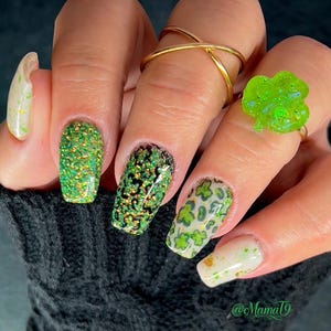 May include: Close-up of a hand with green glitter, shamrock, and gold glitter nail polish. The hand is wearing a gold ring and a green shamrock ring. The caption on the image is "@MamaT9".