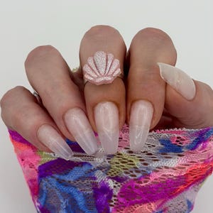 May include: A pink seashell ring with a silver band. The ring is set against a white background. The seashell has a glittery texture. The nails are long and painted with a clear polish.
