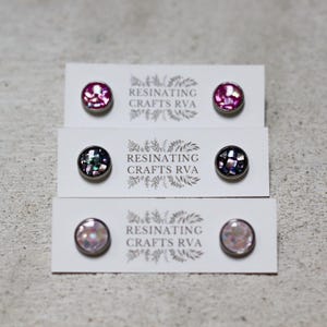 May include: Three pairs of stud earrings with iridescent, mosaic-like surfaces are displayed on white cards. The earrings are in shades of pink, black, and white. Each card is printed with the text "RESINATING CRAFTS RVA" and a floral design.