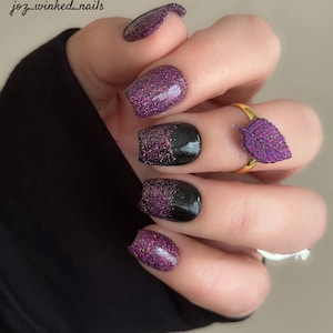 May include: Close-up of a hand with short, square-shaped nails painted with a black and purple glitter gradient. The ring finger has a gold ring with a purple leaf charm.