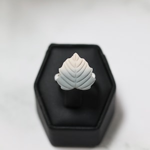 May include: A silver ring featuring a detailed leaf design. The leaf is light gray with subtle shading, transitioning to a pale pink at the top. The ring is displayed on a black hexagonal stand against a white background.