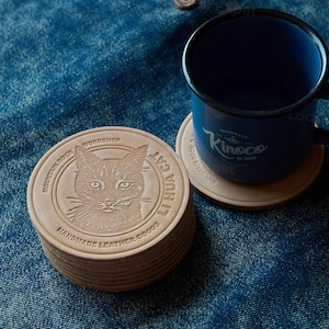 May include: A stack of round, light brown leather coasters with a cat face design and the text "LIHUA CAT" and "HANDMADE LEATHER GOODS". A blue enamel mug sits on a matching coaster. The mug has the text "Kinoco" on it.