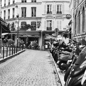 May include: Black and white street scene in Paris, France, featuring cobblestone streets, buildings with awnings, and parked scooters. The image captures a quaint European atmosphere with shops and restaurants.