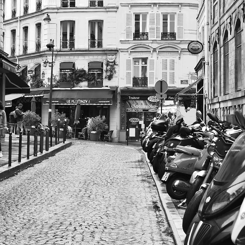 May include: Black and white street scene in Paris, France, featuring cobblestone streets, buildings with awnings, and parked scooters. The image captures a quaint European atmosphere with shops and restaurants.