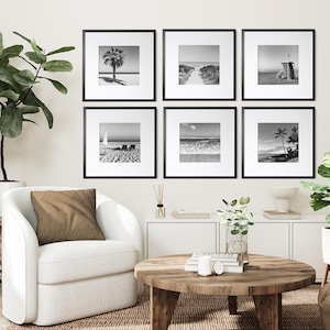 May include: Six framed black and white beach landscape prints arranged on a wall. The images depict scenes of palm trees, ocean waves, and beach chairs. The frames are square with white matting.
