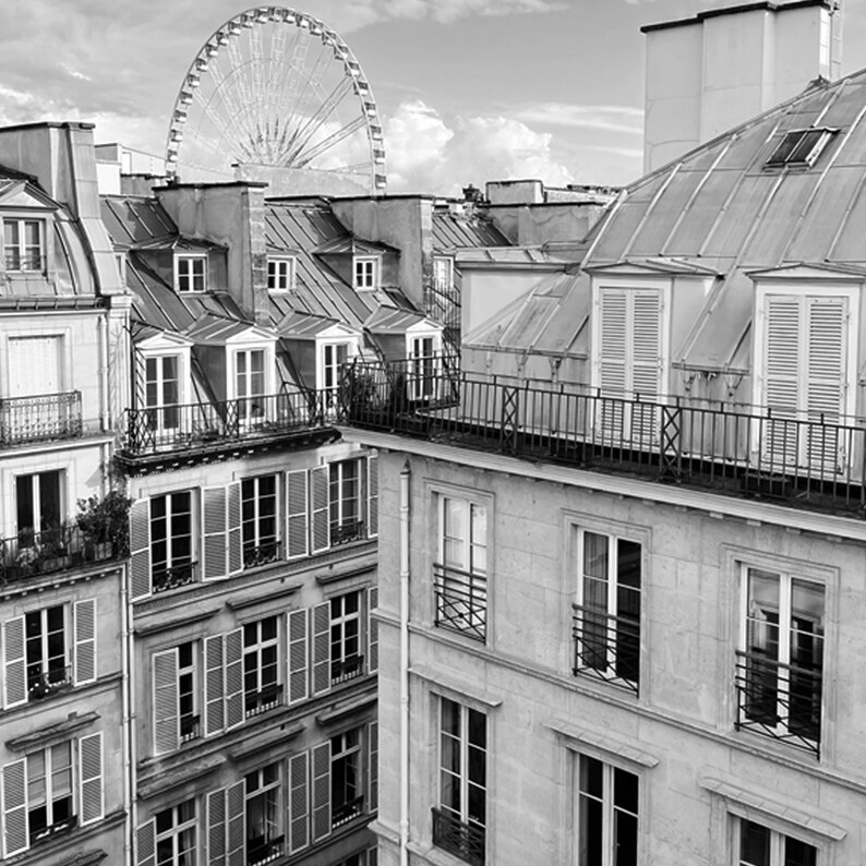 May include: Black and white photograph of Parisian buildings with multiple stories, featuring windows with shutters and balconies. A Ferris wheel is visible in the background, set against a cloudy sky. The image captures the architectural details of the city.