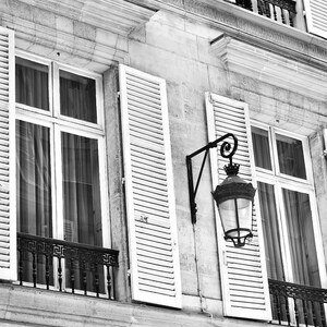 May include: Black and white architectural photograph of a building facade with white shutters, windows, and a decorative wrought-iron balcony. A vintage street lamp is mounted on the wall.