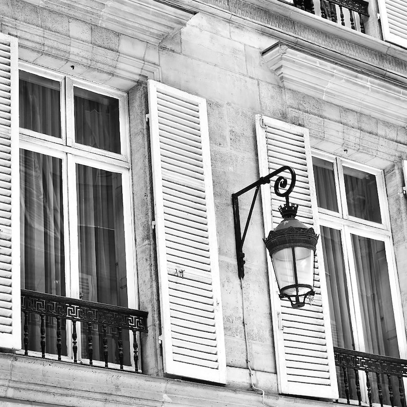 May include: Black and white architectural photograph of a building facade with white shutters, windows, and a decorative wrought-iron balcony. A vintage street lamp is mounted on the wall.