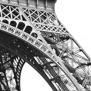 May include: Black and white photograph of a section of the Eiffel Tower. The intricate iron lattice structure is visible, with rivets and crossbeams creating a geometric pattern. Names are inscribed at the top. The image is a close-up, emphasizing the architectural details.