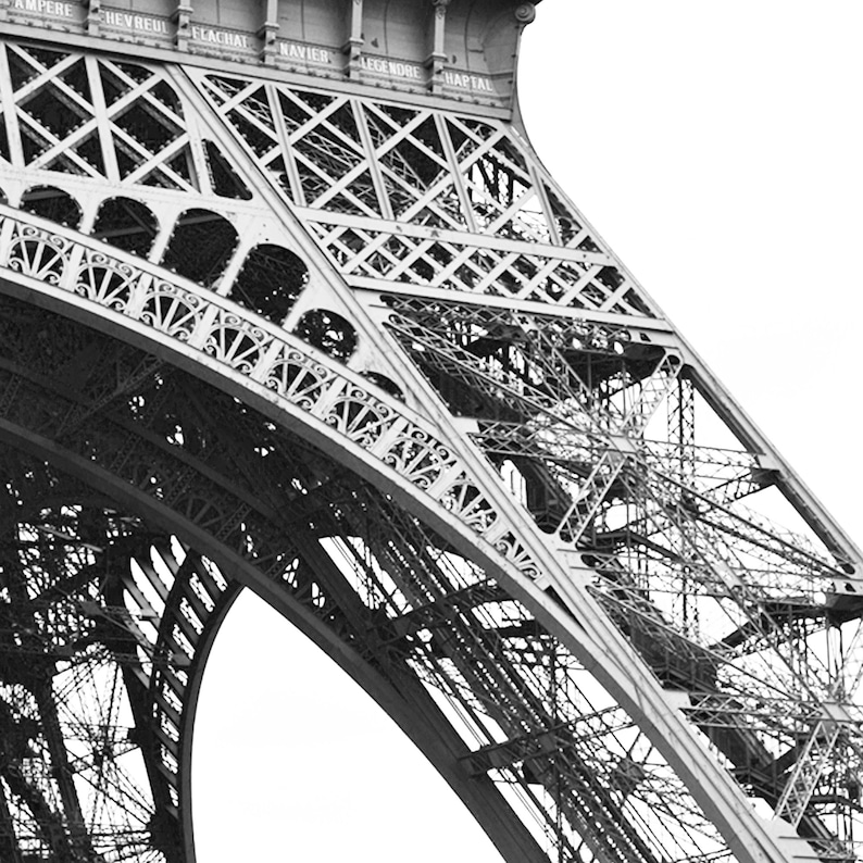 May include: Black and white photograph of a section of the Eiffel Tower. The intricate iron lattice structure is visible, with rivets and crossbeams creating a geometric pattern. Names are inscribed at the top. The image is a close-up, emphasizing the architectural details.