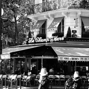 May include: Black and white photograph of a Parisian cafe, "Le Champ de Mars", with outdoor seating. The cafe has awnings, a sign, and a second-story balcony. Tables and chairs are set up outside, and a scooter and bicycles are parked nearby.