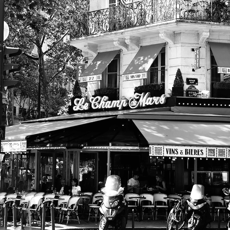 May include: Black and white photograph of a Parisian cafe, "Le Champ de Mars", with outdoor seating. The cafe has awnings, a sign, and a second-story balcony. Tables and chairs are set up outside, and a scooter and bicycles are parked nearby.