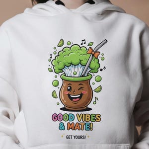 Peut inclure: Sweat à capuche blanc avec un motif coloré d'une tasse de maté souriante avec des feuilles vertes et une paille. Le texte "GOOD VIBES & MATE!" est imprimé en dessous. Le sweat à capuche a une poche avant et une capuche à cordon.