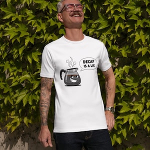 May include: White t-shirt with a cartoon coffee pot graphic and the text "DECAF IS A LIE" in a speech bubble. The coffee pot has a smiling face and steam rising from the spout. The shirt is worn by a person.