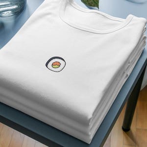 May include: A stack of white t-shirts with a small embroidered sushi roll design on the front. The sushi roll features a black seaweed wrap, white rice, and a salmon filling. The shirts are neatly folded on a blue table.