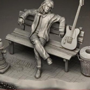 Op de afbeelding: Een grijs, gebeeldhouwd beeldje van een persoon die op een bank zit met een gitaar die tegen de bank leunt. Het beeldje staat op een grijze basis met de woorden "Kurt Cobain" erop gegraveerd.