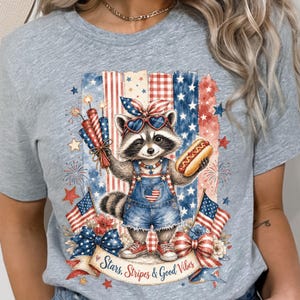 May include: A light gray t-shirt featuring a raccoon wearing sunglasses and denim overalls, holding a hot dog and fireworks. The design includes American flags, stars, and the text "Stars, Stripes & Good Vibes."