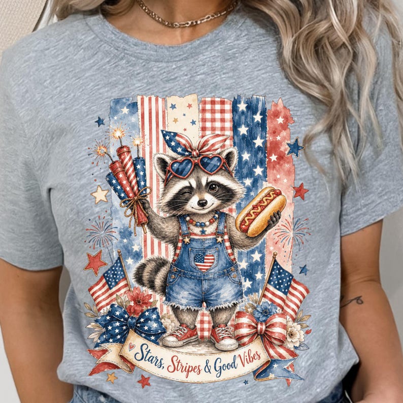 May include: A light gray t-shirt featuring a raccoon wearing sunglasses and denim overalls, holding a hot dog and fireworks. The design includes American flags, stars, and the text "Stars, Stripes & Good Vibes."