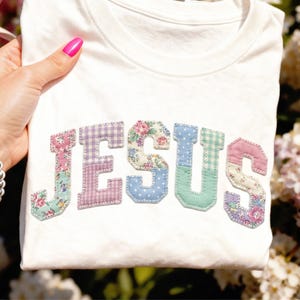 Può includere: Una maglietta bianca con la scritta "JESUS" applicata sul davanti. Le lettere sono in stile patchwork, utilizzando vari tessuti floreali e fantasia in colori pastello. La maglietta è tenuta in mano con smalto rosa.
