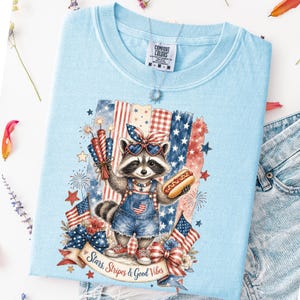 May include: Light blue t-shirt featuring a raccoon wearing heart-shaped sunglasses and denim overalls, holding a hot dog and fireworks. The design includes American flags, stars, and the text "Stars, Stripes & Good Vibes."