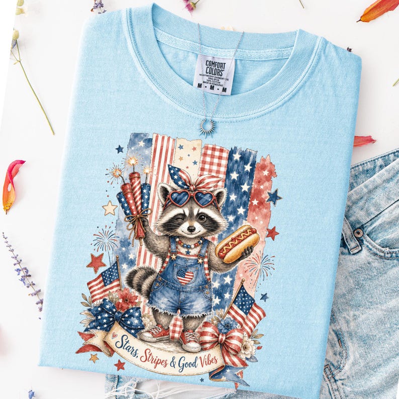 May include: Light blue t-shirt featuring a raccoon wearing heart-shaped sunglasses and denim overalls, holding a hot dog and fireworks. The design includes American flags, stars, and the text "Stars, Stripes & Good Vibes."