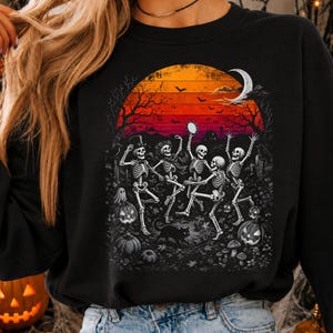 May include: A black sweatshirt featuring a graphic print of four skeletons dancing in a graveyard under a large orange and red sunset. The scene includes a crescent moon, bats, a black cat, jack-o'-lanterns, and trees against a dark background.