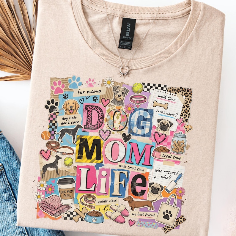 May include: Beige t-shirt with a colorful graphic design. The design includes the words "DOG MOM LIFE" with various dog-themed illustrations, hearts, paw prints, and the phrases "fur mama", "dog hair don't care", "walk time", "treat time", and "cuddle vibes".