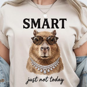 Op de afbeelding: Beige t-shirt met het woord "SMART" in grote zwarte letters boven een capibara met een bril en een diamanten ketting. De zin "just not today" staat eronder in cursief.