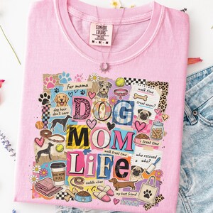 May include: A light pink t-shirt with a colorful graphic that reads "DOG MOM LIFE." The design includes dog illustrations, paw prints, hearts, and the phrases "fur mama," "dog hair don't care," and "my best friend."