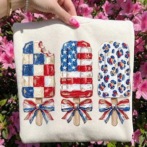 May include: Cream-colored sweatshirt featuring three popsicle designs. One popsicle has a checkered pattern, another depicts the American flag, and the third has a leopard print. Each popsicle has a bow at the base. The background is pink flowers.