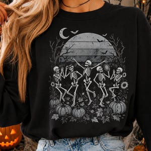 May include: A black long-sleeve sweatshirt featuring a graphic print of five skeletons dancing in a field of pumpkins and sunflowers under a large moon and bare trees. The design includes silhouettes of flying bats against the night sky.