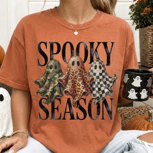 May include: An orange short-sleeve t-shirt featuring the black text SPOOKY SEASON and three ghost illustrations. The left ghost has a green camouflage pattern, the center ghost has a brown leopard print, and the right ghost has a black and white checkerboard pattern.