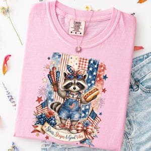 May include: Pink Comfort Colors t-shirt featuring a patriotic raccoon wearing sunglasses and holding a hot dog and fireworks. The raccoon is set against a backdrop of American flags and fireworks. The text "Stars, Stripes & Good Vibes" is on a banner.