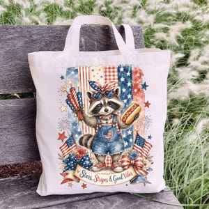 May include: A white tote bag featuring a raccoon wearing sunglasses and denim overalls, holding a hot dog and fireworks. The design incorporates red, white, and blue elements, including stars, stripes, and American flags. Text reads "Stars, Stripes & Good Vibes."