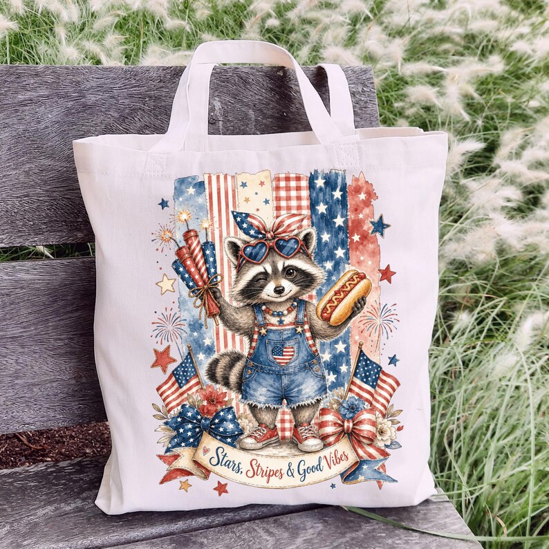 May include: A white tote bag featuring a raccoon wearing sunglasses and denim overalls, holding a hot dog and fireworks. The design incorporates red, white, and blue elements, including stars, stripes, and American flags. Text reads "Stars, Stripes & Good Vibes."