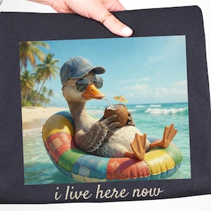 May include: A graphic print on a black background features a duck wearing sunglasses and a hat, relaxing in a colorful inflatable ring in the ocean. The duck is holding a coconut drink with a straw. The text "i live here now" is at the bottom.