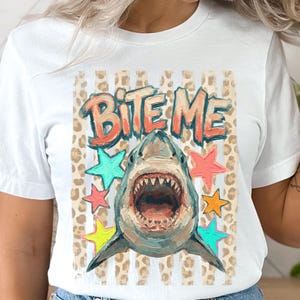 Può includere: Una t-shirt bianca con una stampa grafica di uno squalo con la bocca aperta. Sopra lo squalo, il testo BITE ME è scritto in arancione. Il design include uno sfondo a strisce verticali beige con stampa leopardata e stelle colorate.