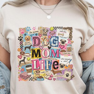 May include: Beige t-shirt with a colorful graphic design featuring the words "DOG MOM LIFE" and various dog-related illustrations. The design includes paw prints, hearts, dog breeds, and the text "fur mama".