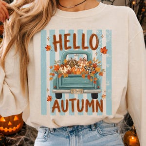 Puede incluir: Una sudadera color crema con un gráfico de una camioneta vintage azul claro llena de calabazas estampadas y hojas de otoño. El texto HELLO AUTUMN está impreso en marrón encima y debajo de la camioneta sobre rayas azules.