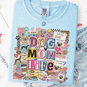 May include: Light blue Comfort Colors t-shirt with a colorful collage design. The design includes the words "DOG MOM LIFE" with dog-related graphics and phrases like "dog hair don't care" and "walk time". A silver necklace is visible.