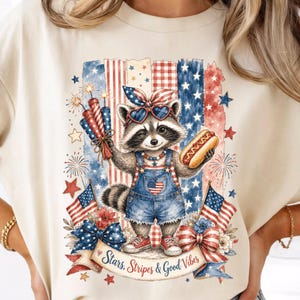 May include: Cream-colored t-shirt featuring a patriotic raccoon wearing sunglasses and denim overalls, holding fireworks and a hot dog. The design includes American flags, stars, and the text "Stars, Stripes & Good Vibes."
