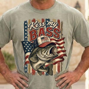 Op de afbeelding: Lichtgroen t-shirt met een afbeelding van een basvis met een hoed met een Amerikaans vlagontwerp. De tekst "Kiss my BASS" staat boven de vis, met een rood, wit en blauw Amerikaanse vlag op de achtergrond.