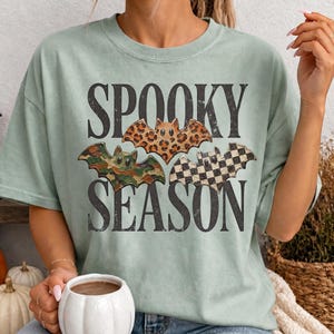 May include: A light green t-shirt featuring the text SPOOKY SEASON in black capital letters. Between the words are three illustrated bats: one with a brown leopard print, one with a green camouflage pattern, and one with a black and white checkerboard.