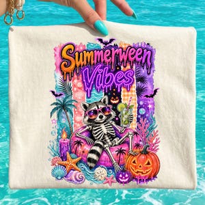 Peut inclure: Sac fourre-tout crème avec un squelette de raton laveur portant des lunettes de soleil et tenant un cocktail, se prélassant sur un anneau gonflable rose. Le sac porte le texte "Summerween Vibes" avec des graphismes sur le thème d'Halloween et de la plage.