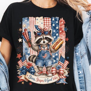 May include: Black t-shirt featuring a raccoon wearing heart-shaped sunglasses and denim overalls, holding fireworks and a hot dog. The design includes American flags, stars, and the text "Stars, Stripes & Good Vibes."